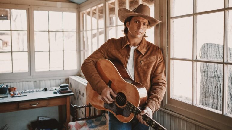 Lukas Nelson holds acoustic guitar in rustic interior, promotional photo for Palace Theatre Albany show
