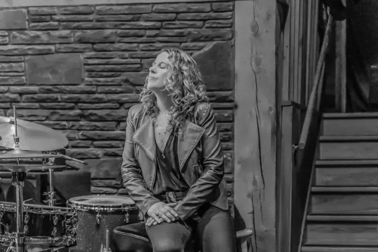 Amy Helm seated on a stool beside a drum kit at Levon Helm Studios, eyes closed, with a stone wall behind her