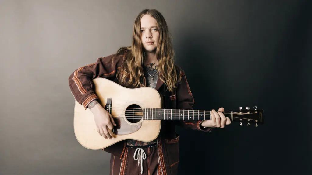 Billy Strings holds an acoustic guitar in a professional press photo