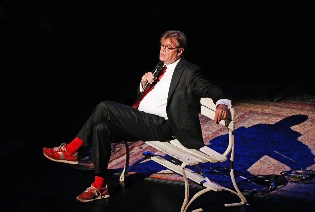Garrison Keillor performs on stage seated on a bench, holding a microphone