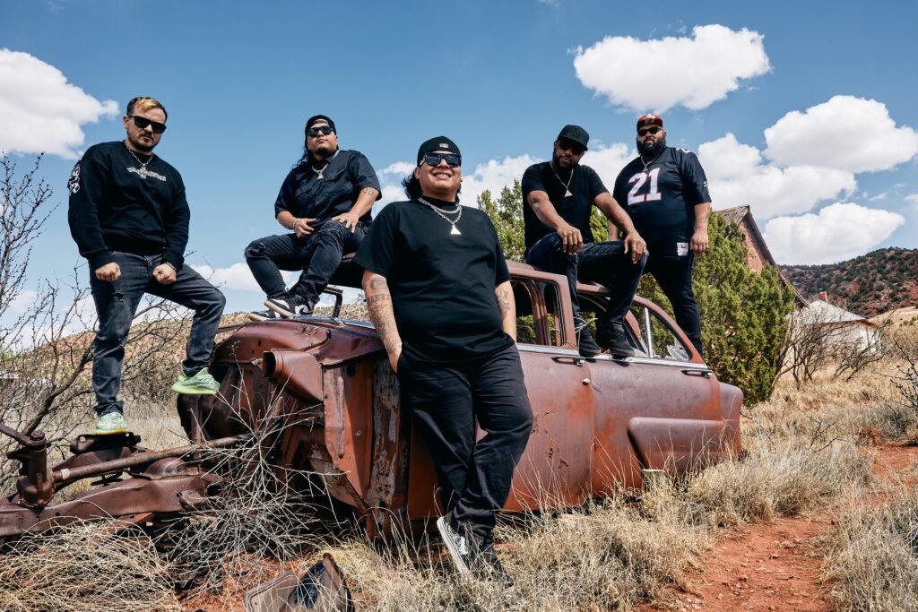 Giovannie & The Hired Guns pose around a rusted vintage car in a Southwest desert landscape