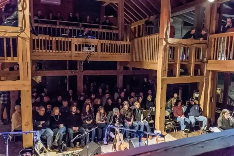 Wide interior view of Levon Helm Studios in Woodstock, NY, showing vaulted wooden ceilings, a wraparound wooden balcony, and a packed audience facing the stage during a Midnight Ramble