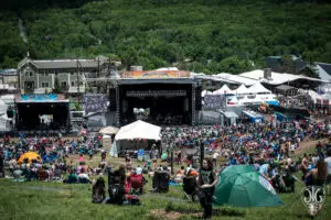 Wide view of Mountain Jam festival at Hunter Mountain Ski Resort showing the main stage with Mountain Jam signage, two video screens, festival tents, and a crowd spread across the green ski slope with Catskill forest in the background