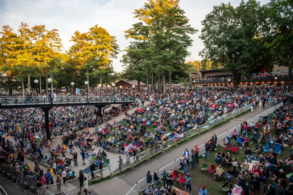 Wide elevated view of the lawn at the Saratoga Performing Arts Center taken from the back, audience seated on chairs and blankets across the slope, pavilion roof visible in the distance through the trees