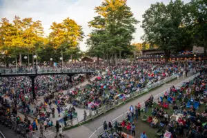 Wide elevated view of the lawn at the Saratoga Performing Arts Center taken from the back, audience seated on chairs and blankets across the slope, pavilion roof visible in the distance through the trees