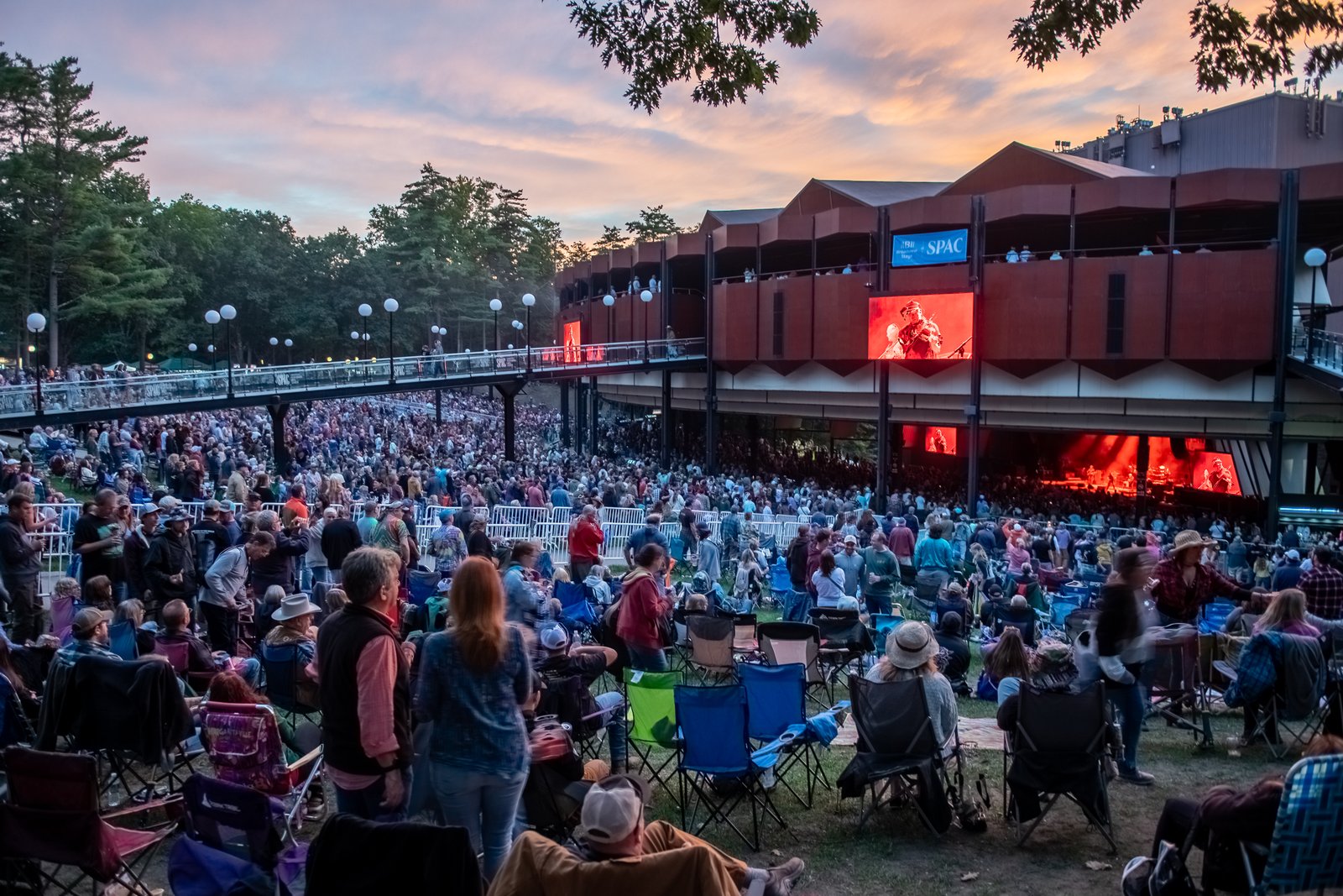 View from the front of the SPAC lawn looking toward the pavilion, the cantilevered roof of the open-air structure projecting outward, the venue's video screen lit and an artist visible mid-set