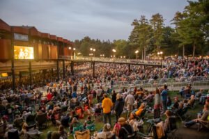 Saratoga Performing Arts Center pavilion at dusk with the lawn full of audience members on chairs and blankets, venue lights on, big screen showing the performance, pine trees and twilight-blue sky behind the structure