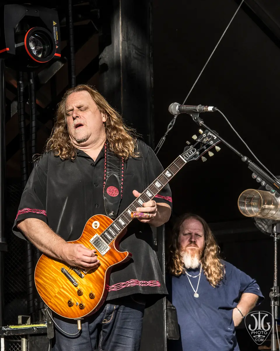 Warren Haynes performing at Mountain Jam at Hunter Mountain June 2013 with eyes closed mid-solo on a cherry sunburst Gibson Les Paul; longtime guitar tech Brian Farmer visible in blue shirt in the background