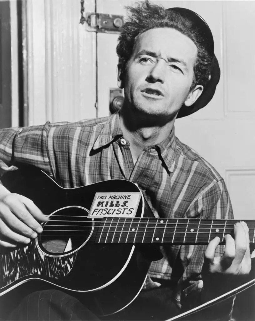 Woody Guthrie with guitar bearing "This Machine Kills Fascists" sticker, 1943