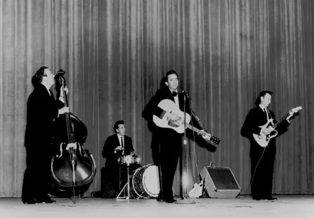 Black-and-white performance photograph of Johnny Cash playing acoustic guitar at center microphone, flanked by Marshall Grant on upright bass at left, W.S. Holland on drums at center-rear, and Luther Perkins on electric guitar at right; 1963.