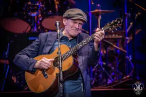 James Taylor performing onstage at Saratoga Performing Arts Center, wearing a flat cap and dark blazer, eyes closed in concentration, playing acoustic guitar under warm spotlights against a blue and purple backdrop.