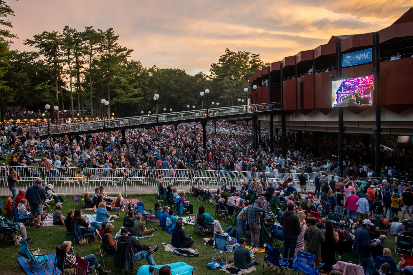 Wide twilight view of the Saratoga Performing Arts Center amphitheater with the lawn crowd, pedestrian bridge, and tiered pavilion lit by golden-hour sky, video screen showing the performance.