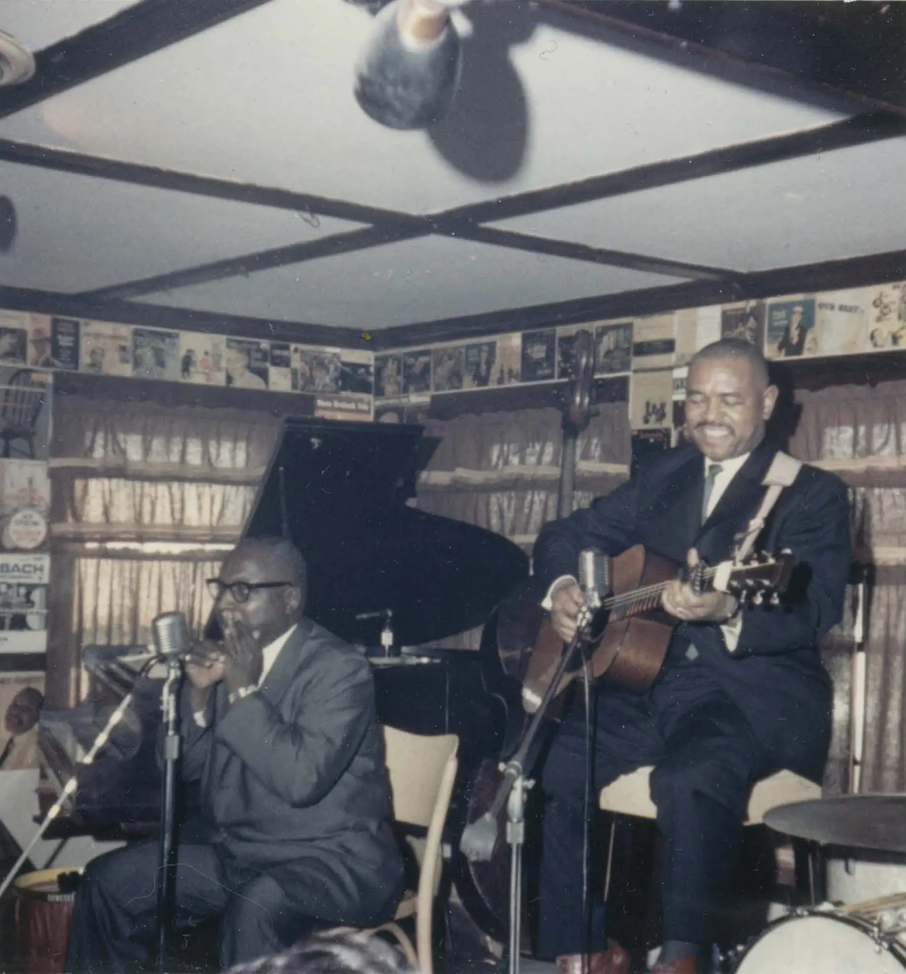 Color photograph of blues musicians Sonny Terry and Brownie McGhee performing on a small stage, February 1964; Terry plays harmonica into a vintage microphone, McGhee plays acoustic guitar and smiles.
