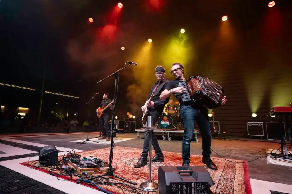 BoDeans performing live on stage with guitar, accordion, and full band under colorful stage lighting
