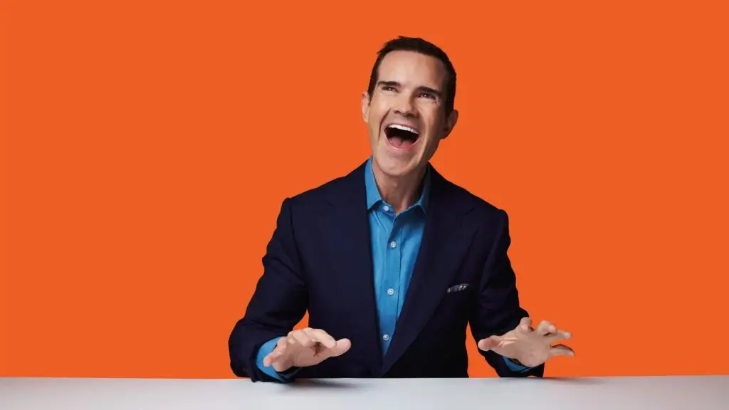 Jimmy Carr performing stand-up comedy, laughing with hands spread wide against orange background