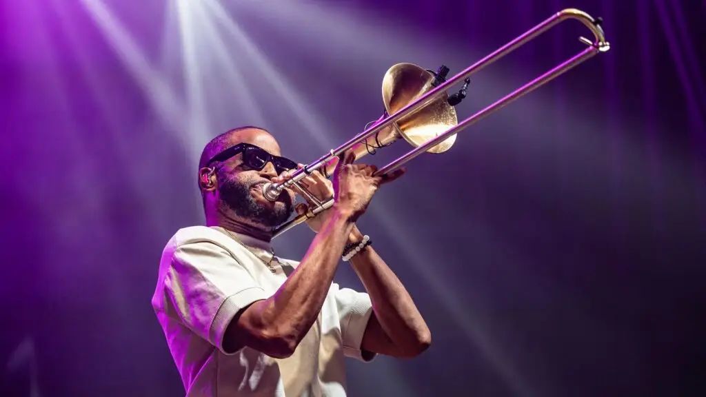 Trombone Shorty performing live on stage, playing trombone under dramatic purple stage lights