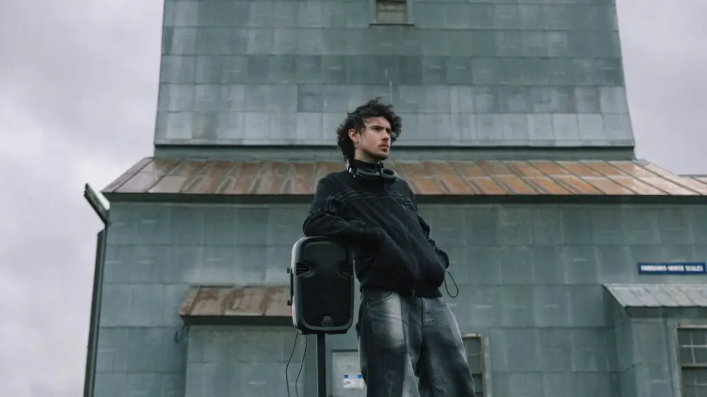 Quadeca stands outdoors near a portable speaker with headphones around his neck, 2026 press photo