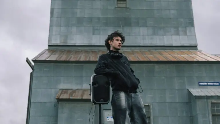 Quadeca stands outdoors near a portable speaker with headphones around his neck, 2026 press photo