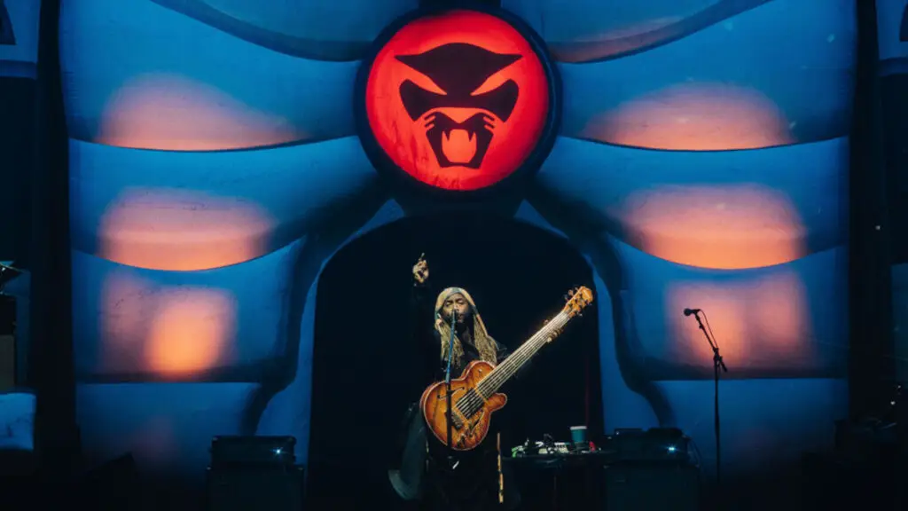 Thundercat performing live on stage, playing his signature 6-string bass beneath his panther logo backdrop