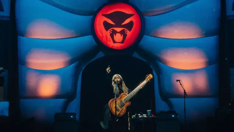 Thundercat performing live on stage, playing his signature 6-string bass beneath his panther logo backdrop