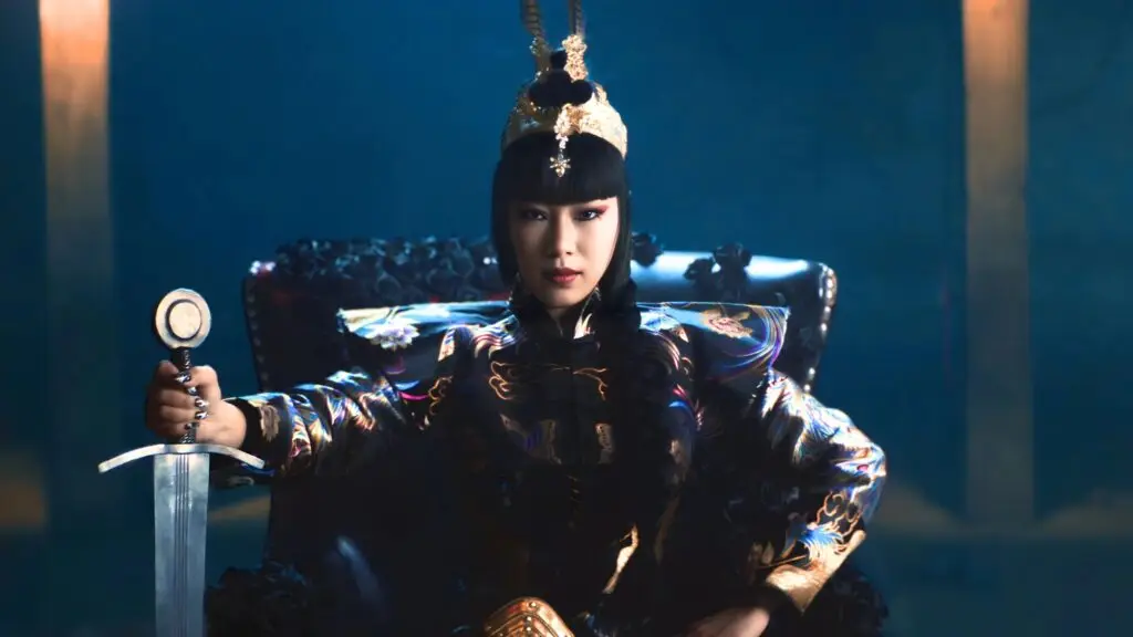 NINI seated in traditional Chinese regalia with ornate embroidered robe and golden crown, holding a sword