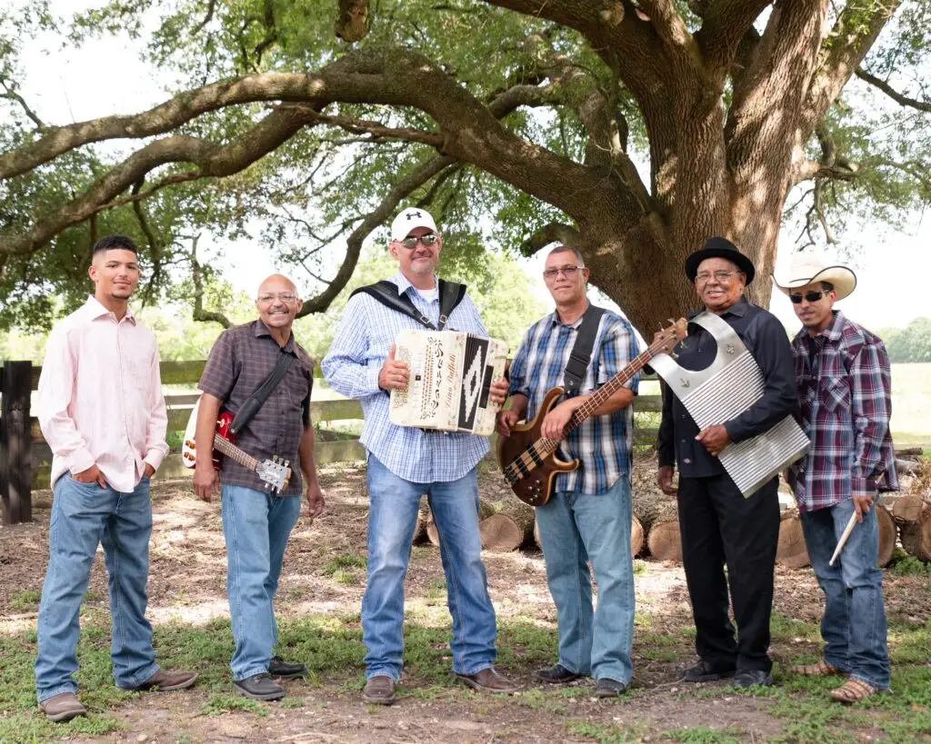 Horace Trahan and the Ossun Express band posing outdoors under a large oak tree with accordion, bass, and scrubboard