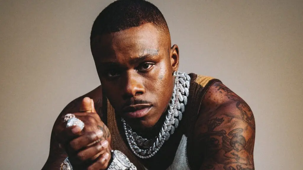 DaBaby performing live, leaning forward with intensity, diamond chains and sleeve tattoos visible