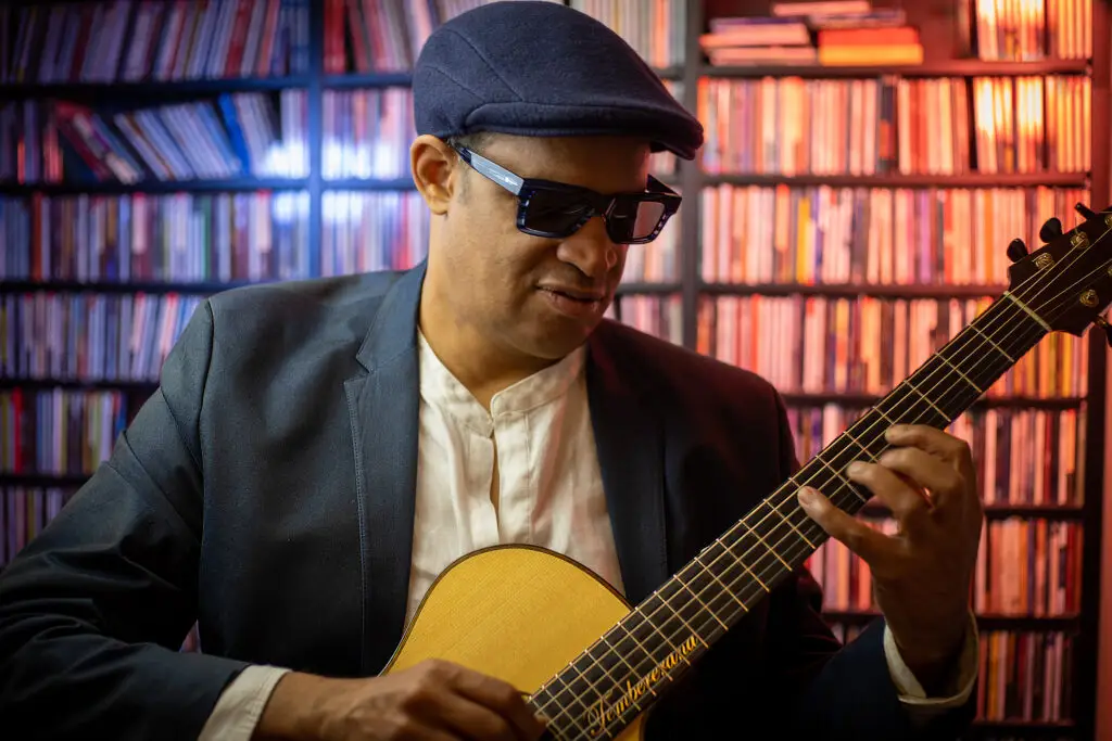 Raul Midón playing acoustic guitar, wearing dark sunglasses and a flat cap, seated in front of shelves of CDs