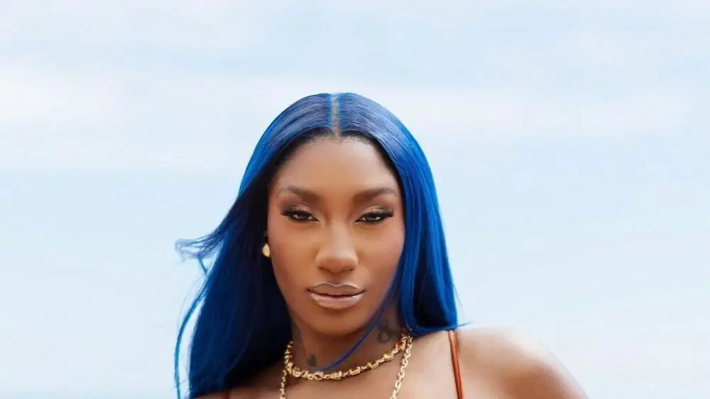Aya Nakamura with electric blue hair and gold chain necklaces, outdoor coastal photoshoot