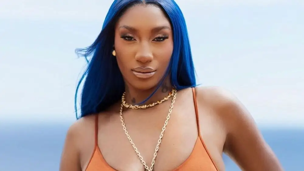 Aya Nakamura with electric blue hair and orange bodysuit, outdoor coastal setting