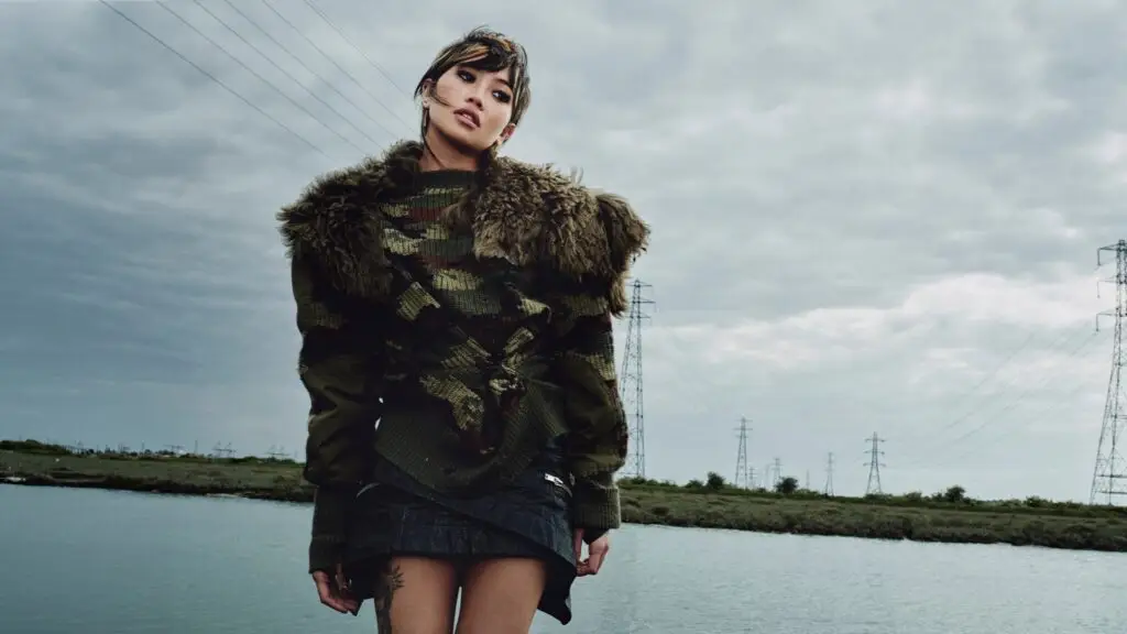 beabadoobee stands outdoors near electrical pylons in a fur-trimmed camo sweater, Pylon album era