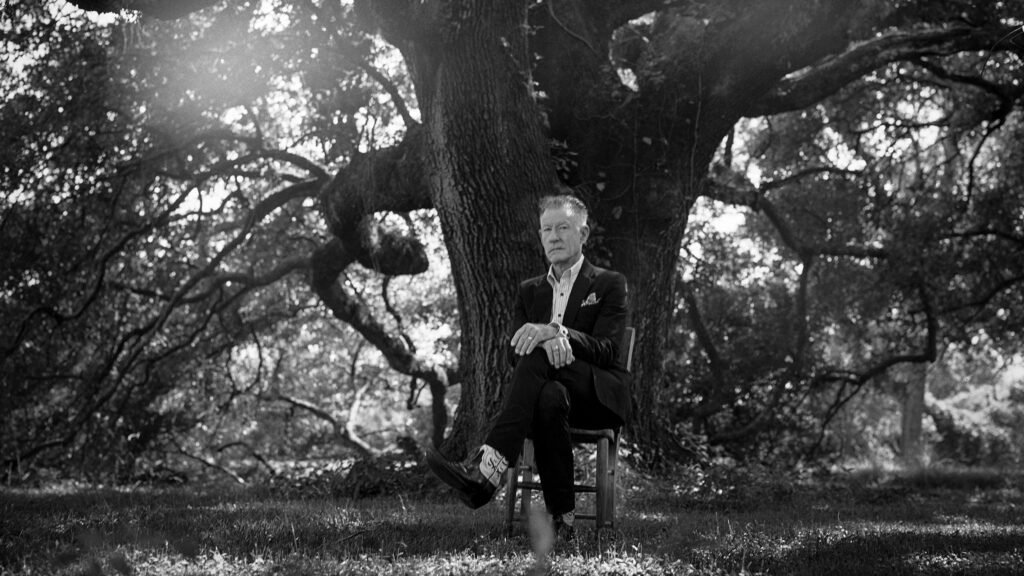 Lyle Lovett seated beneath a live oak tree in a dark suit and cowboy boots, press photo