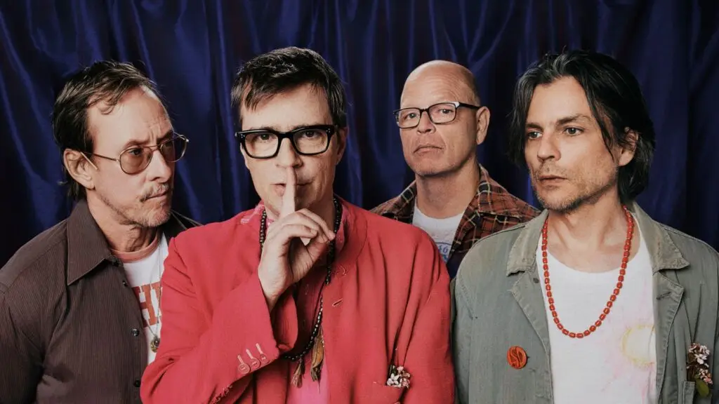 Weezer band press photo featuring Rivers Cuomo (center, red blazer) with Brian Bell, Pat Wilson, and Scott Shriner