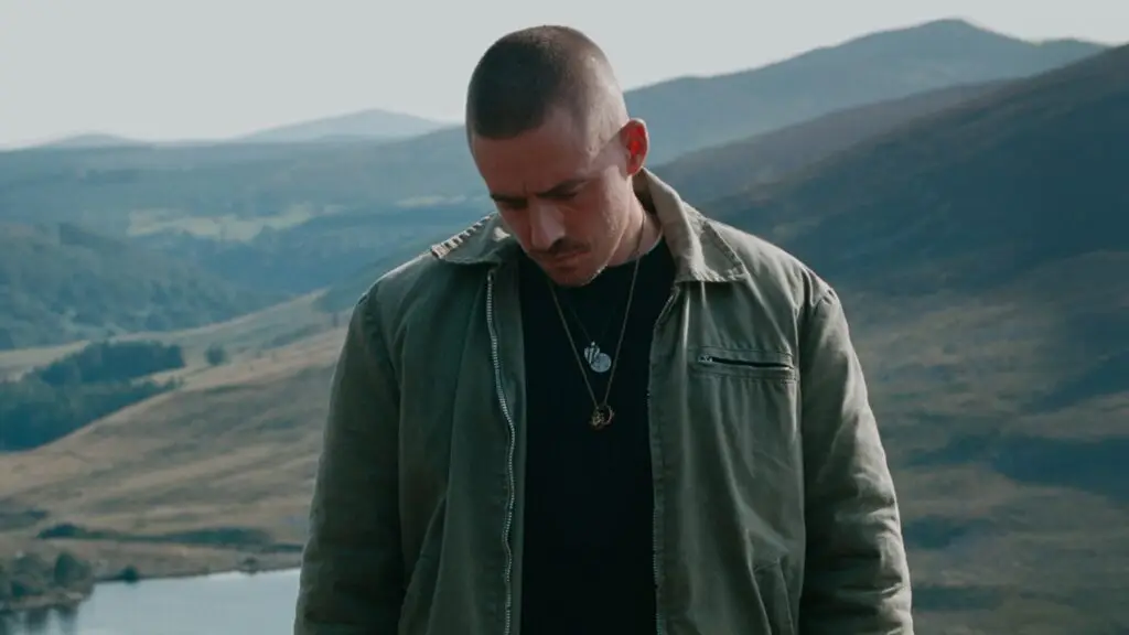 Dermot Kennedy stands in an Irish highland landscape, olive jacket and chain necklaces, Weight of the Woods era