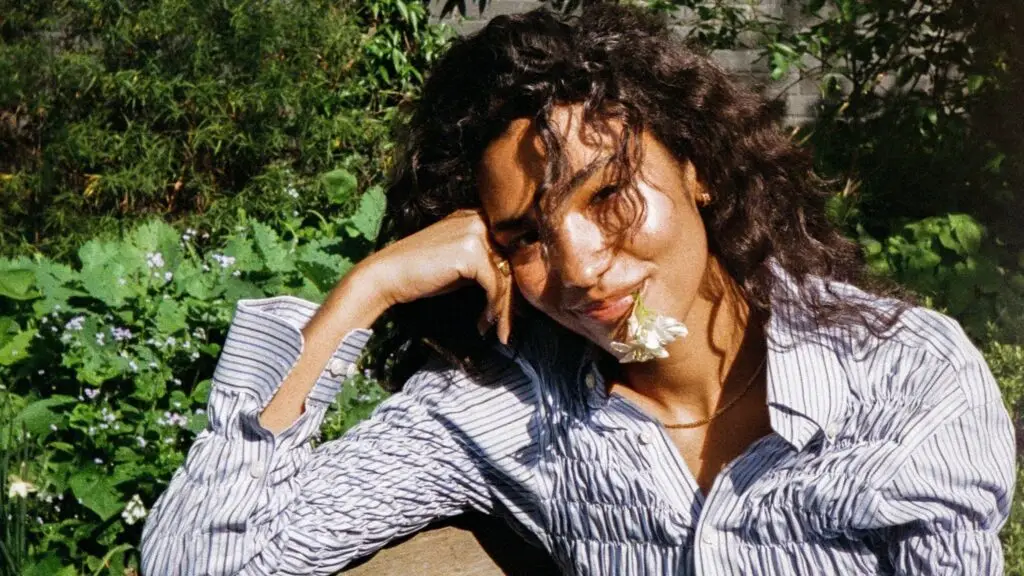 Olivia Dean in a sun-dappled garden, holding a white flower, press photo for The Art of Loving Tour