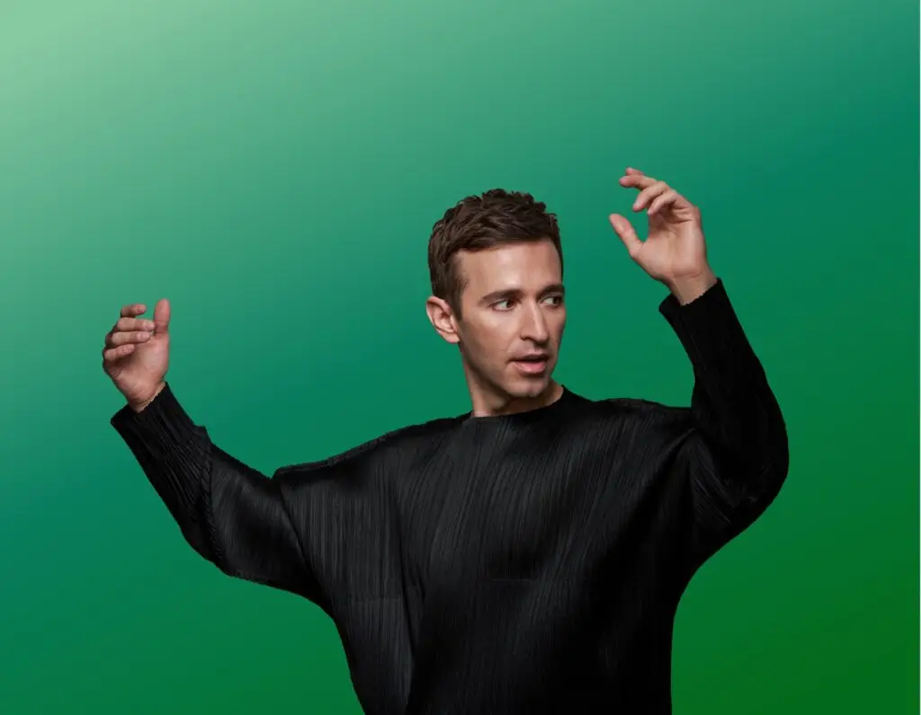 Anthony Roth Costanzo, countertenor, in an expressive performing pose with both hands raised against green studio backdrop