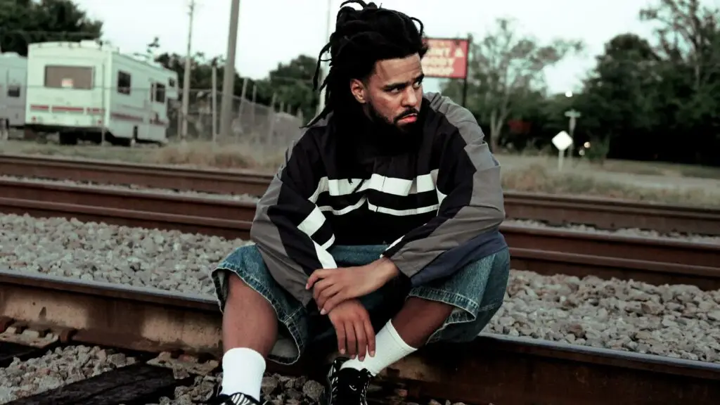 J. Cole seated on railroad tracks in a suburban setting, dreadlocks, The Fall Off Tour 2026