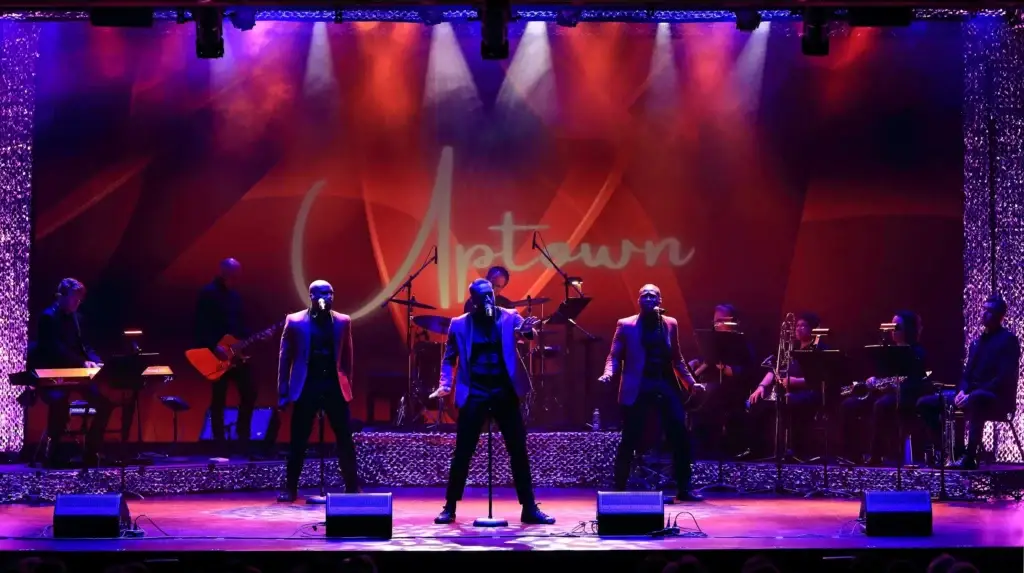 Uptown performing live on stage, three vocalists in matching blazers with full band