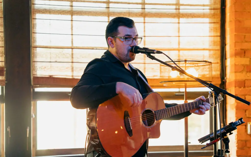 Alex Goettel of Palisade performing live at mic with acoustic guitar, warm indoor Rochester venue
