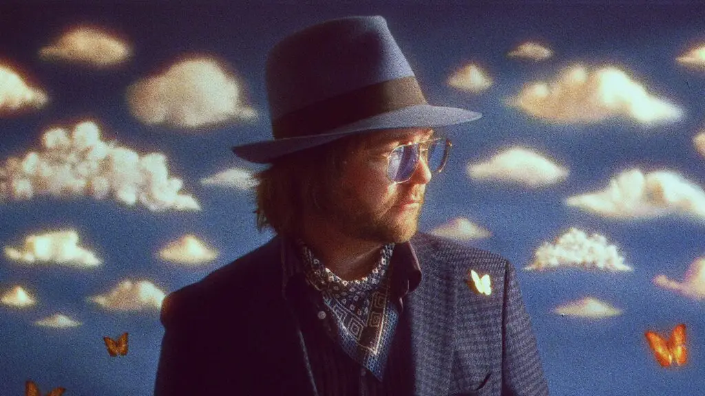 Aaron Lee Tasjan in fedora and tweed jacket, clouds and butterflies backdrop