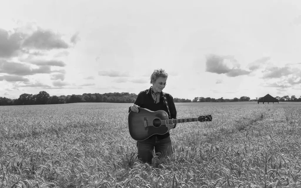 Mary Gauthier with acoustic guitar in a wheat field, black-and-white 2026 press photo