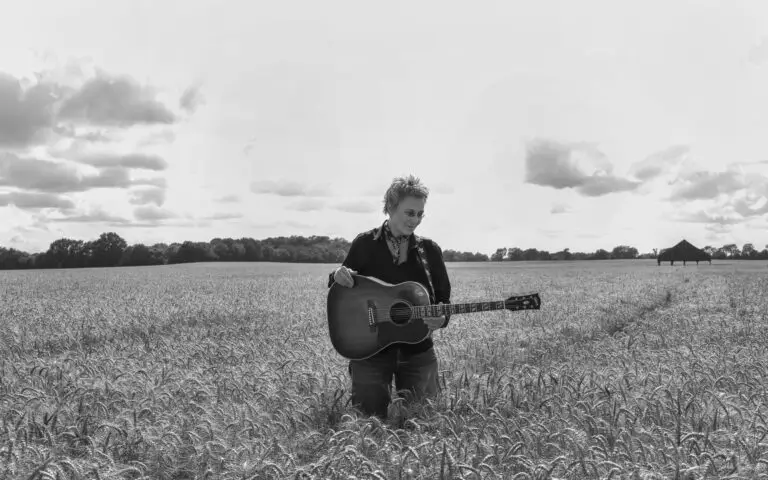Mary Gauthier with acoustic guitar in a wheat field, black-and-white 2026 press photo