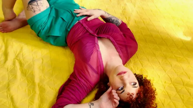 Carsie Blanton in a vibrant artistic press photo, curly auburn hair and tattoo sleeves, reclining on bright yellow fabric