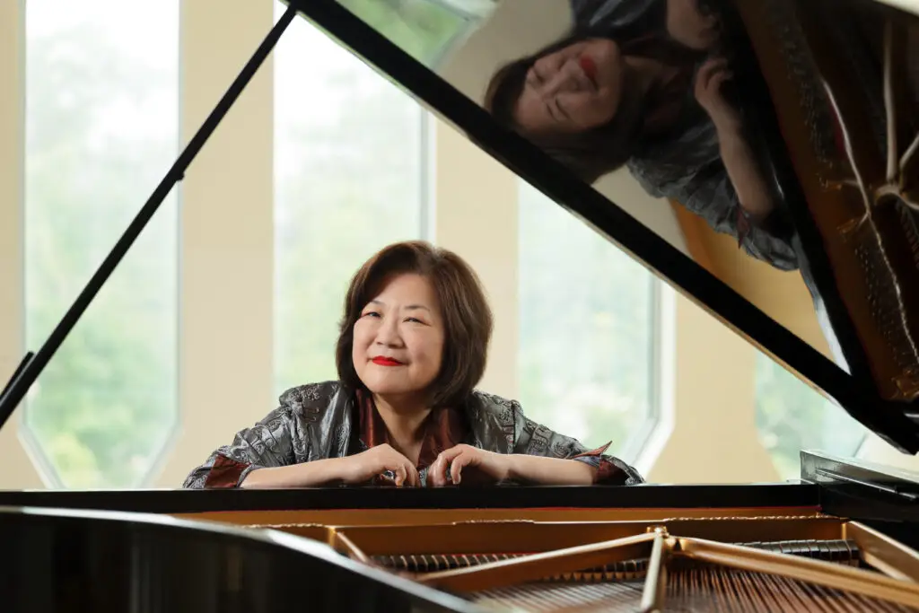 Angela Cheng leaning on a grand piano, smiling, with her reflection in the piano lid