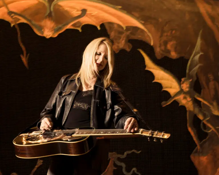 Cindy Cashdollar playing Dobro at an outdoor festival stage, blonde hair, black jacket
