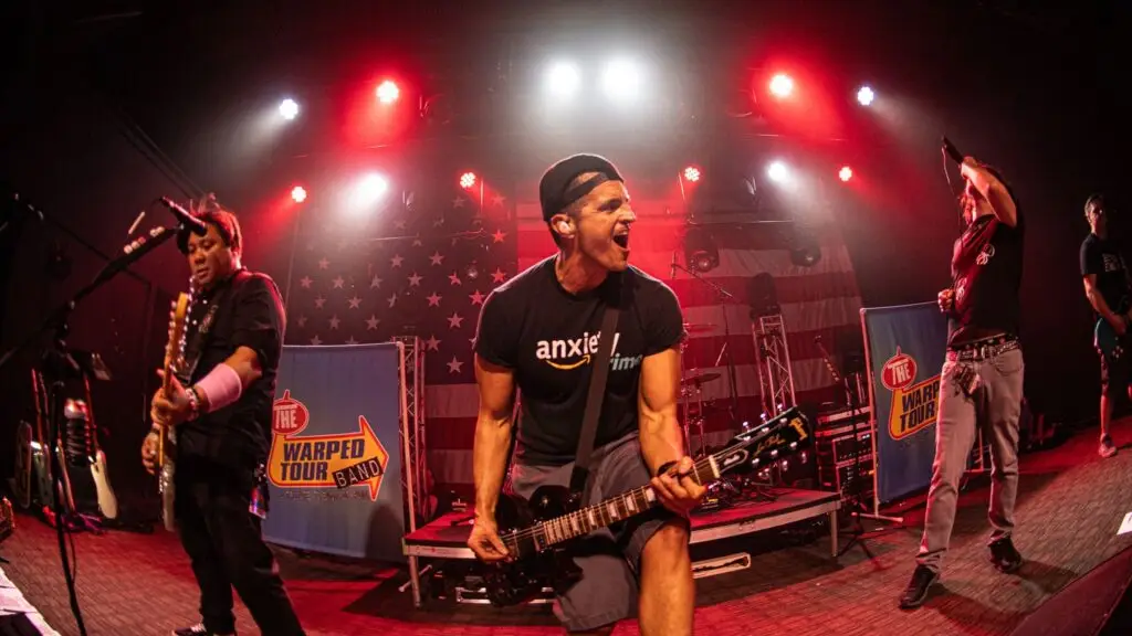 The Warped Tour Band performing live on stage with band logo banners and American flag backdrop