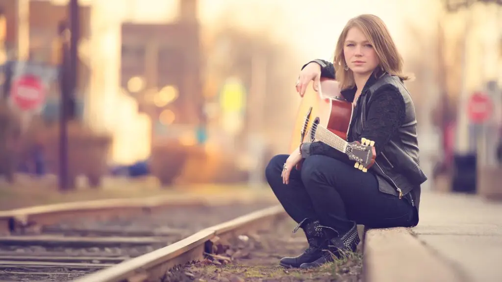 Crystal Bowersox with acoustic guitar seated on railroad tracks, outdoor press photo
