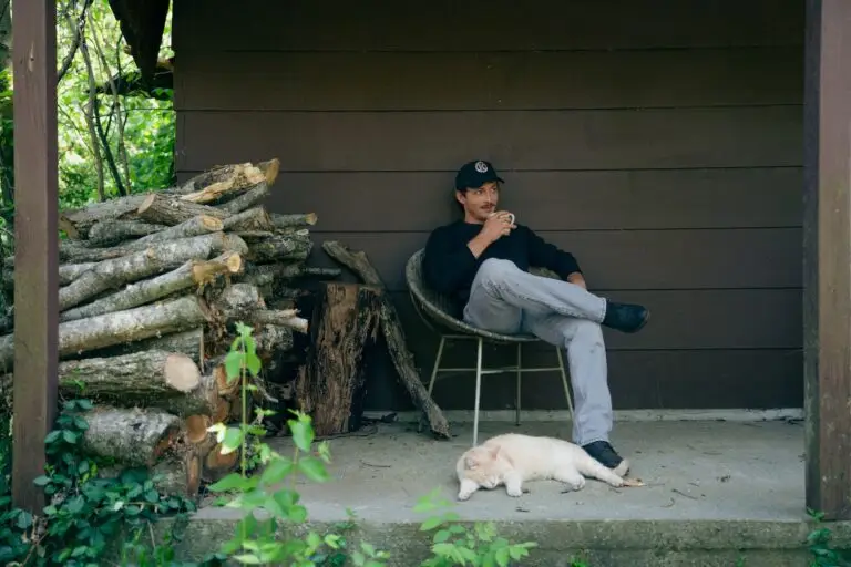 Hunter Flynn seated on a cabin porch holding a coffee mug, white cat resting at his feet