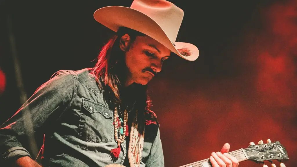 Duane Betts performing live, focused on guitar, wearing a cream cowboy hat and beaded necklaces