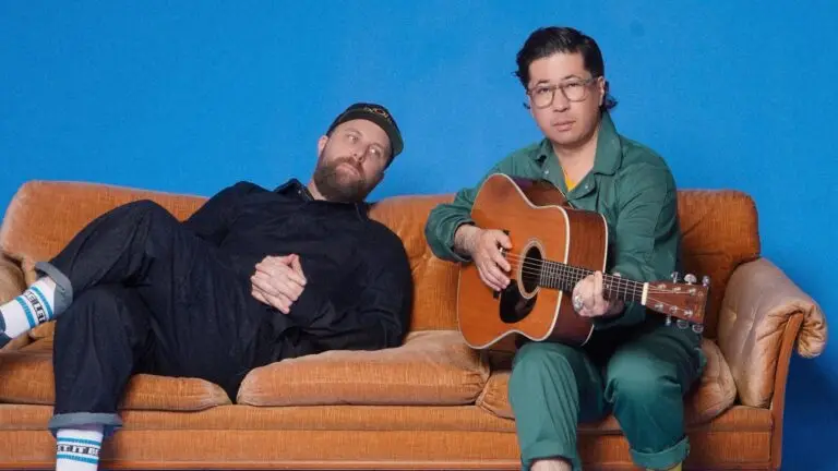 Choir! Choir! Choir! co-directors Daveed Goldman and Nobu Adilman on couch with acoustic guitar, blue studio backdrop